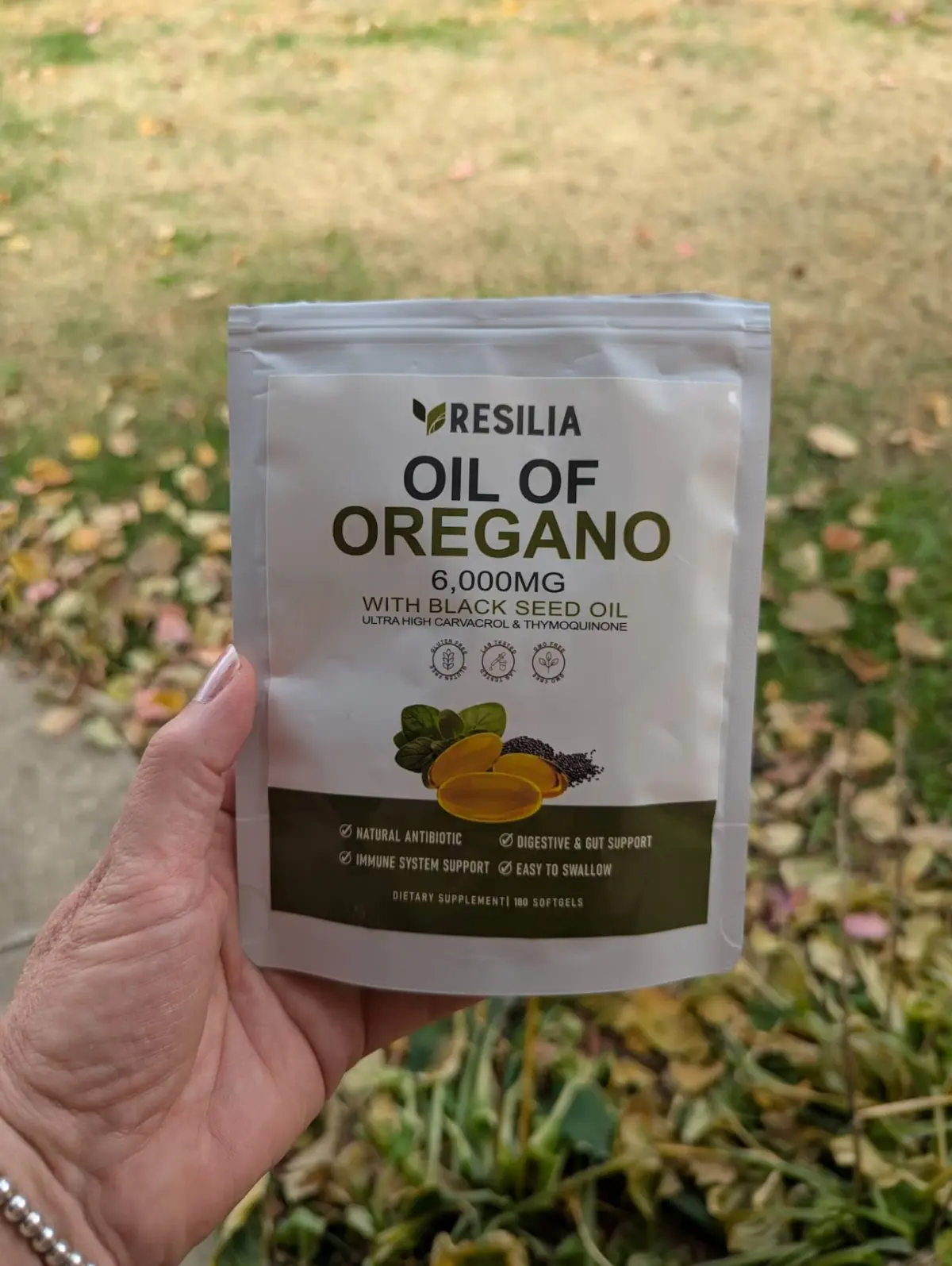 Hand holding a pouch of Resilia Oil of Oregano 6,000mg with black seed oil softgels, outdoors on a grassy lawn.