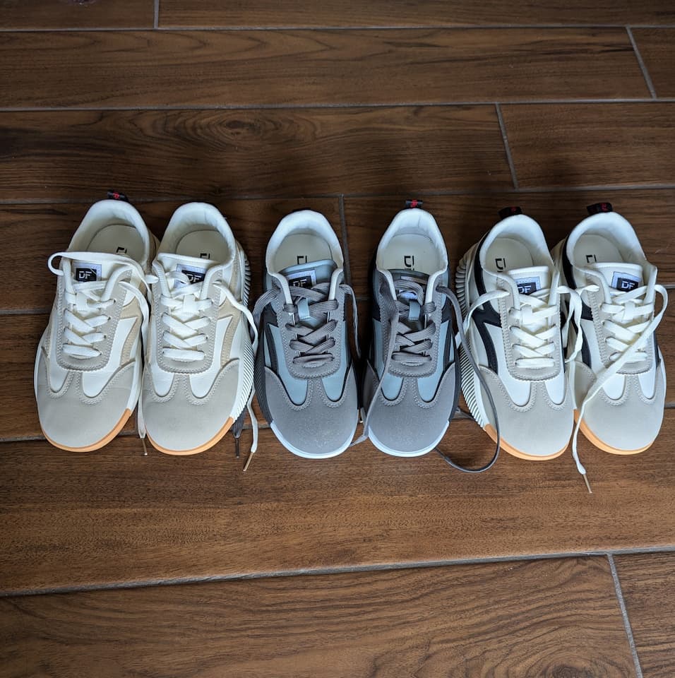 Three pairs of De Florance sneakers in cream, gray, and black lined up on a hardwood floor.