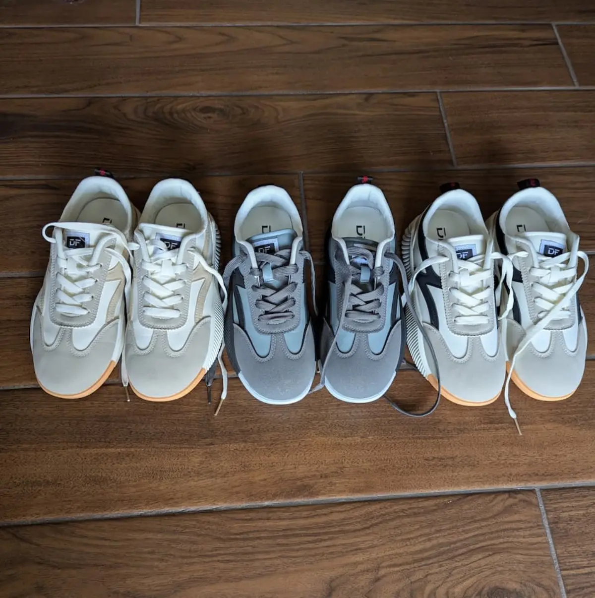 Three pairs of De Florance sneakers in cream, gray, and black lined up on a hardwood floor.