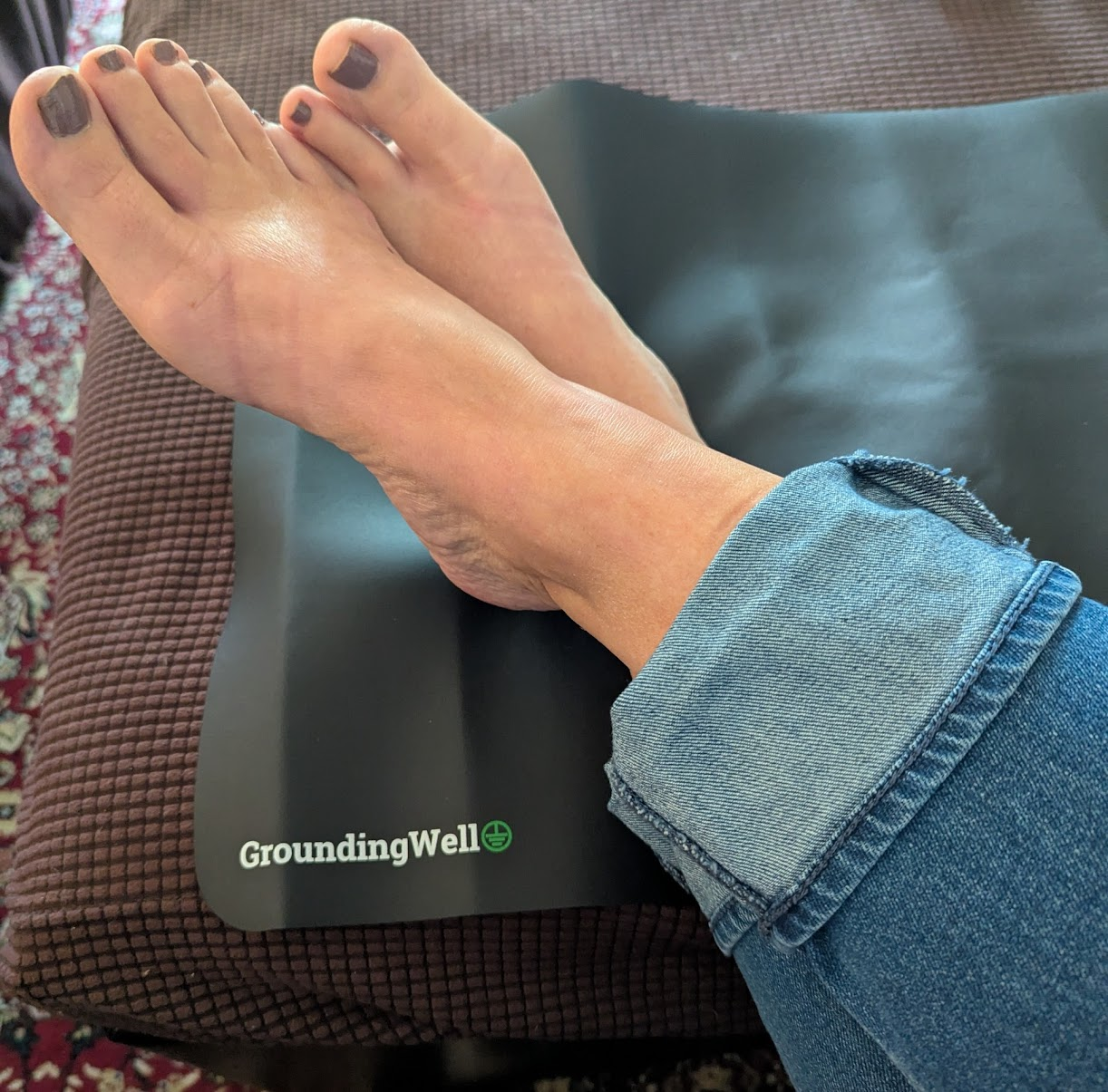 Bare feet resting on a Grounding Well wellness mat with cuffed blue jeans, styled for an indoor grounding routine.