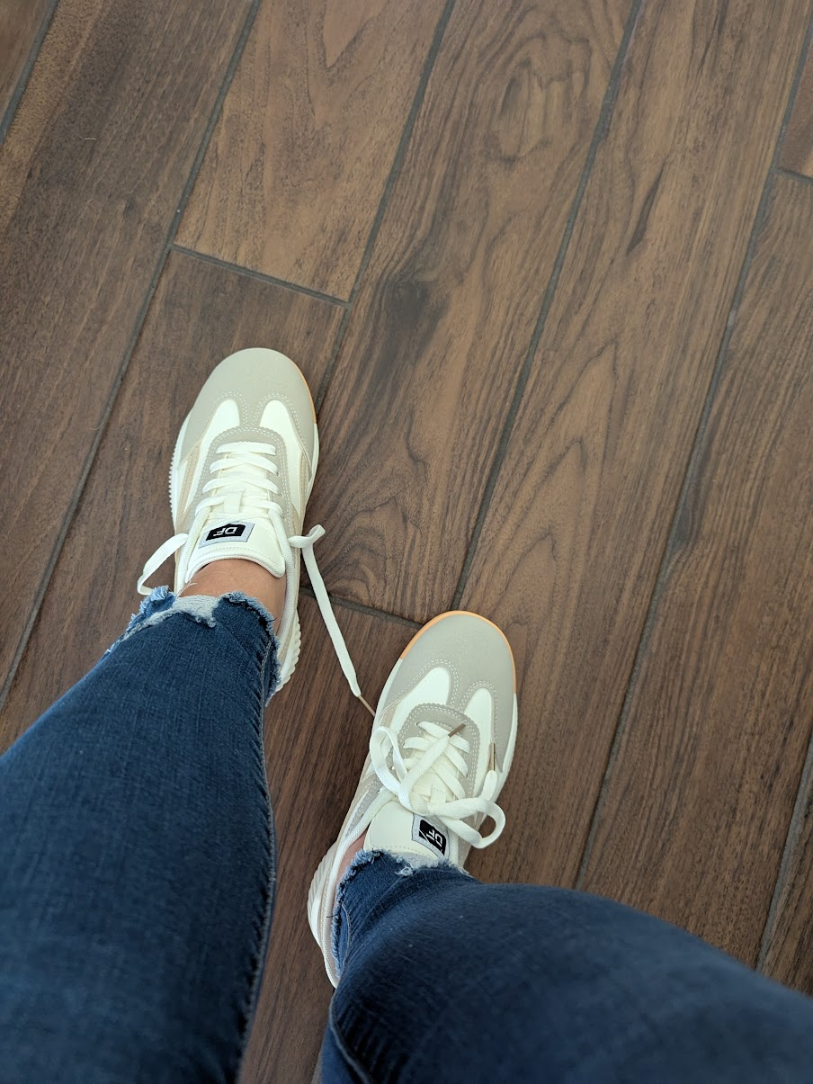 Top-down view of feet wearing white orthopedic tennis shoes paired with dark jeans on a wood floor.