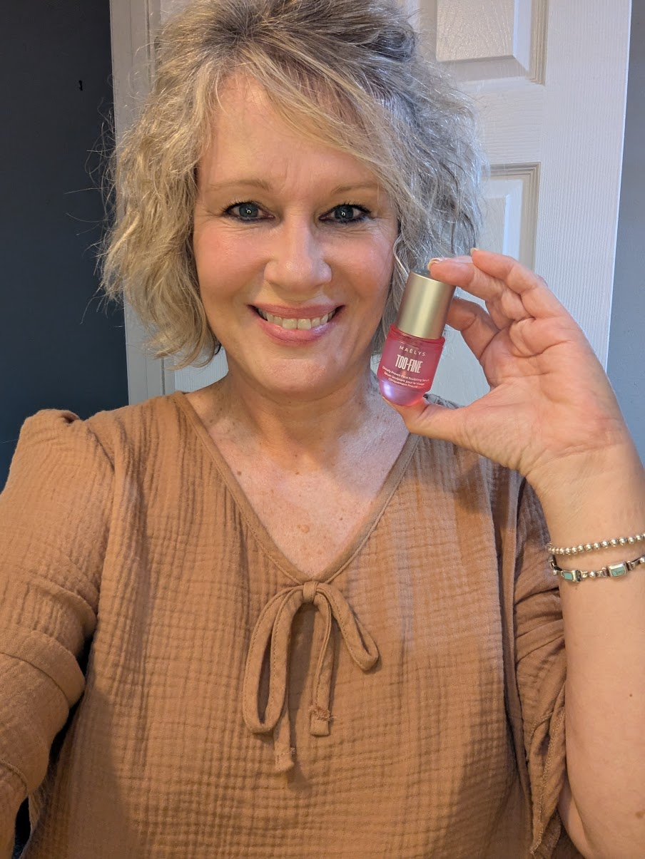 Alica Rutherford smiling and holding up a favorite skincare product, wearing a tan blouse.